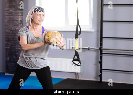 Angenehme Tätigkeit. Positive fröhliche senior Frau hält einen Ball und training mit ihm während er in der Turnhalle Stockfoto