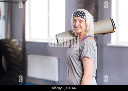 Aktiven Lebensstil. Positiv im Alter von schönen Frau hält eine Yoga-Matte und in eine positive Stimmung während der Zeit im Fitnessstudio verbringen Stockfoto