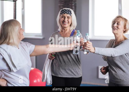 Für gesunden Lebensstil. Fröhlich glücklich Rentnerinnen halten Flaschen mit Wasser und Klirren mit ihnen beim Ausruhen nach dem Fitness-training Stockfoto