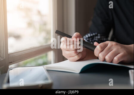 Frau Hand mit Stift beim Schreiben auf kleinen Notebook neben Fenster. Freier Journalist in Heimarbeit Konzept. Stockfoto