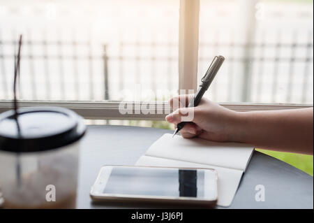 Frau Hand mit Stift beim Schreiben auf kleinen Notebook mit Smartphone auf Tisch neben Fenster. Freier Journalist in Heimarbeit Konzept. Stockfoto