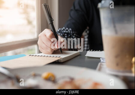 Frau Hand mit Stift beim Schreiben auf kleinen Notebook neben Fenster. Freier Journalist in Heimarbeit Konzept. Stockfoto