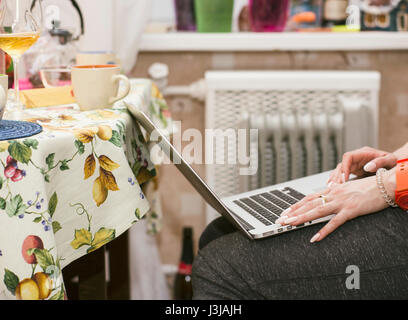 Weibliche Hände Tippen auf einen Laptop auf den Knien, Küche Interieur in den unscharfen Hintergrund platziert. Gefilterte Schuss Stockfoto