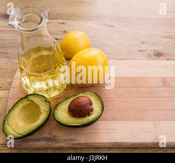 Avocado-Öl in Karaffe mit Avocado in halbe und zwei ganze Zitronen neben Dekanter auf ein Schneidebrett auf Holz Hintergrund schneiden. Über dem Winkel Ansicht, Zimmer Stockfoto