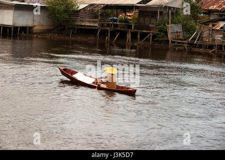 Ein asiatischer Mann seine kleine Ruderboot mit einem kleinen Paddel auf einem braunen farbigen Flussufer am Nachmittag Singkawang. Stockfoto