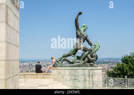 Zwei Personen suchen in ihr Smartphone s anstelle der Ansicht vom Gellertberg in Budapest beim sitzen neben einer Statue eines Mannes, der einen Drachen tötet. Stockfoto