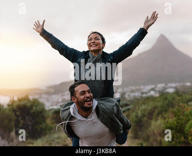 Glücklich junger Mann seine Freundin mit ihrem erhobenen Armen tragen. Paar genießt sich in Landschaft. Stockfoto