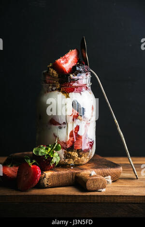 Joghurt-Hafer-Müsli mit Erdbeeren, Maulbeeren, Honig und Minze Blätter in Longdrinkglas Glas auf schwarzem Hintergrund Stockfoto