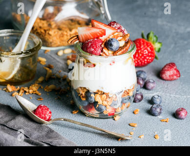 Joghurt-Hafer-Müsli mit frischen Beeren, Nüssen, Honig und Minze im Glas Glas auf grauem Beton strukturiertem Hintergrund Stockfoto