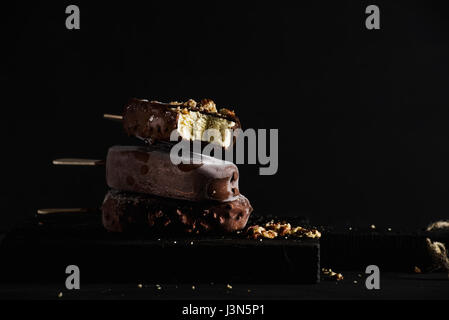 Stapel von Schokolade getauchte Eis am Stiel mit abgebrochenen Muttern auf dunklen Holzbrett auf schwarzem Hintergrund. Stockfoto