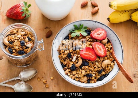 Müsli, Milch und Obst. Leckeres und gesundes Frühstück: Schüssel Müsli mit frischen Erdbeeren, Krug Milch, Mandeln, Bananen und Löffel auf Holz Stockfoto