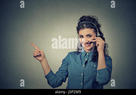 Closeup Portrait schöne, liebenswerte lächelnde Kundenvertreter mit Handy Kopfhörer zeigt auf textfreiraum isoliert auf graue Wand Hinterg Stockfoto