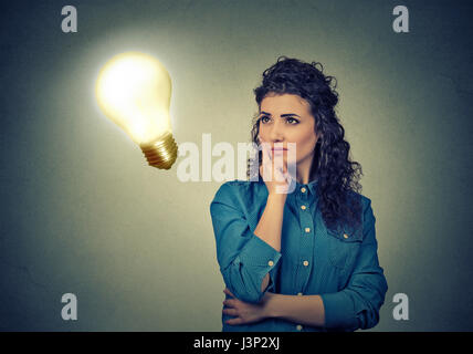 Closeup schönes Mädchen denkt blickte zu helle Glühbirne auf graue Wand Hintergrund isoliert. Konzept, Idee, Bildung und Menschen. Gesichtsausdruck Stockfoto