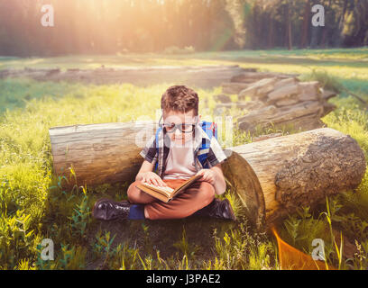 Kleiner Junge mit Schulranzen Lesen eines Buches in Yoga pose, grünen Natur auf Hintergrund. Junge Schüler in Gläsern mit Rucksack und Lehrbuch. Kind mit scho Stockfoto