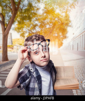 Kleiner Junge in Gläsern liest ein Buch, Stadtstraße auf Hintergrund. Junge Schüler mit Rucksack und Lehrbuch Stockfoto