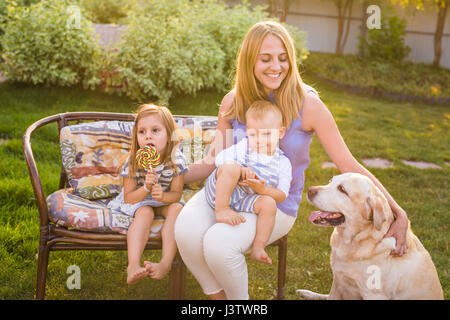 Mutter und ihre Kinder entspannen im Garten mit Hund. Glückliche Familie mit ihrem Labrador Retriever Hund an einem sonnigen Tag spielen Stockfoto