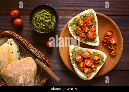 Olivenbrot Roll Hälften mit Basilikum-Pesto bestreichen und garniert mit gebratenen Kirschtomaten-Hälften, serviert auf Holzplatte, overhead auf dunklen wo fotografiert Stockfoto