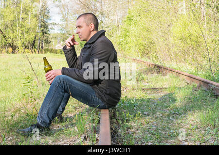 Einsamer Mann mit einer Zigarette und einer Flasche Bier auf verrosteten Schienen sitzen Stockfoto