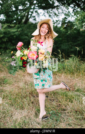 Blumenstrauß, Menschen, Blumen Urlaub, Geschenk und Blumenarrangement Konzept - junge schöne Frau im eleganten Hut posiert mit Blumenstrauß, weiblich in Flip-flops und kokett lächelnd steht auf einem Bein. Stockfoto