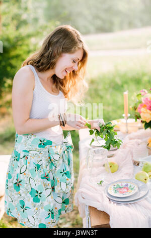 Picknick, Menschen, Essen, Sommer, Urlaub Konzept - junge schöne lächelnde Frau bereiten Minze, Limonade in Glas Flagon auf Tisch rund um festliche Platten, Birne Obst, Bouquet von Blumen, Kerzen. Stockfoto