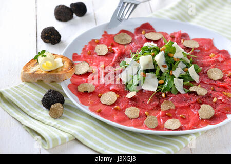 Köstliches Rindfleisch-Carpaccio mit in Scheiben geschnittenen schwarzen Trüffeln, Rucola-Salat mit Parmesan und gerösteten Pinienkernen, serviert mit einer Scheibe geröstetes baguette Stockfoto