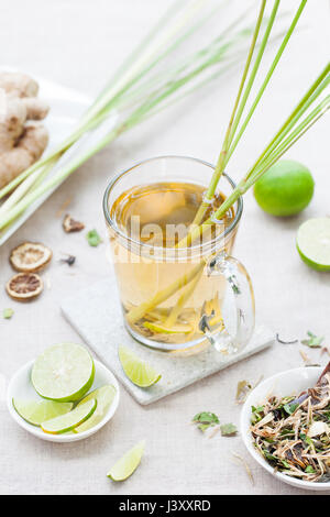 Grün-Kräutertee mit Zitronengras und Ingwer in Glasschale mit frischen Limetten auf textilen Untergrund. Stockfoto