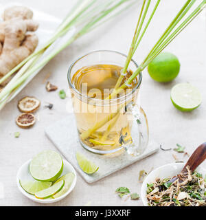 Grün-Kräutertee mit Zitronengras und Ingwer in Glasschale mit frischen Limetten auf textilen Untergrund. Stockfoto