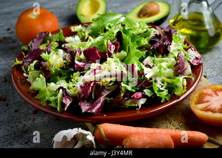 Nahaufnahme eines Steingut Platte mit einem Salat-Mix und einige Zutaten für einen Salat, wie Tomaten, Avocado, Karotten und Olivenöl auf einem grauen bereiten Stockfoto