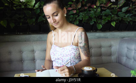 Junge Prety Frau mit Tattoo in ihrem Tagebuch schreiben Tagebuch. Kaukasische Modell mit langen Haaren. Tasse Kaffee und Smartphone auf den Tisch, Stockfoto