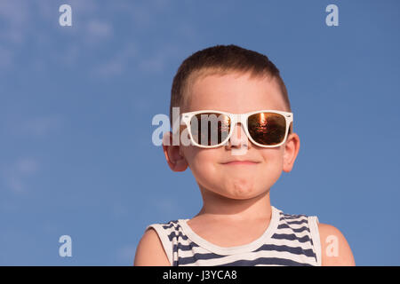 froh, dass kleine Junge tragen weiße Sonnenbrille und Matrosenhemd Streifen auf blauem Himmelshintergrund Stockfoto