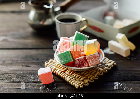 Verschiedene Farbe Turkish Delight Lokum mit Puderzucker auf kleine Teller. Tasse Kaffee und Cezve auf Hintergrund Stockfoto