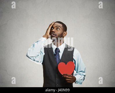 Closeup Portrait junge traurig verwechselt corporate Mann, hält rotes Herz in seinen Händen, zu weinen, Blick nach oben isoliert grau, schwarzer Hintergrund. Negative Stockfoto