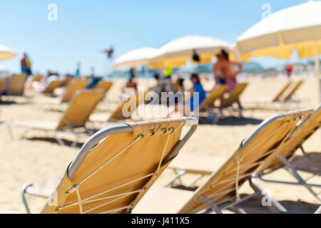 Nahaufnahme von einigen beigen Sonnenliegen und einige Beige Schirme in einem Strand im Mittelmeer, mit vielen unkenntlich Menschen Sonnenbaden in den Rücken Stockfoto