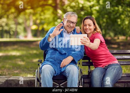 fröhliche Tochter mit ihrem behinderten Vater im Rollstuhl mit Hilfe einer digitalen-Tablets, sprechen und Lächeln während Sie verbringen Zeit im park Stockfoto