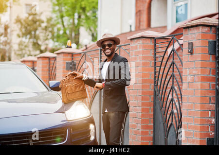 Stilvolle schwarze Mann an mit Hut, Brille tragen Anzug mit Handtasche gegen Luxus-Auto. Reichen afrikanischen amerikanischen Geschäftsmann. Schauen Sie sich Uhren. Stockfoto