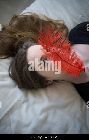 Schöne junge Frau im Bett versteckt Gesicht mit roter Feder zur Festlegung Stockfoto