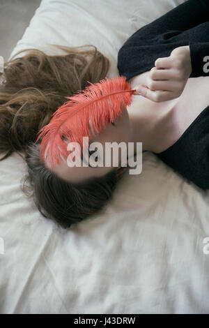 Schöne junge Frau im Bett versteckt Gesicht mit roter Feder zur Festlegung Stockfoto