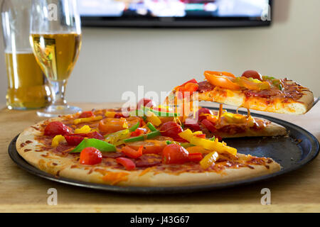 Gemüse Pizza auf einem Tisch mit Bier vor einem Fernseher. Eine Scheibe wird genommen. Stockfoto