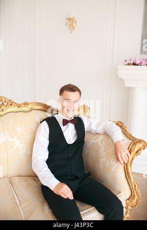 Bräutigam sitzt auf dem Sofa warten auf die Braut am Tag seiner Hochzeit. bei Hochzeit Smoking lächelnd und Braut warten. Elegante Mann im schwarzen Kostüm und b Stockfoto