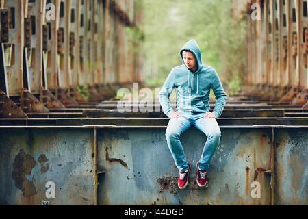 Trauriger Mann sitzt auf der verlassenen rostigen Brücke. Konzept für Traurigkeit, Einsamkeit und vieles mehr. Stockfoto