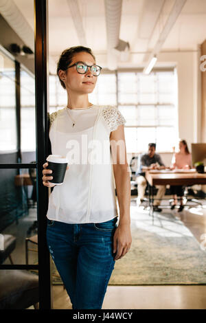 Porträt der schönen jungen Frau, die in Büro-Tür mit Kaffee stehe. Weibliche Führungskraft mit Kaffeepause mit Kollegen im Hintergrund arbeitet. Stockfoto