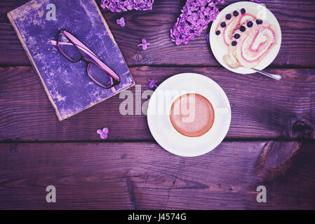 Kaffee in eine weiße Tasse mit einer Untertasse und ein Keks-Kuchen auf einem Holztisch, daneben ist ein altes Buch und Brille, eine Ansicht von oben, ein Vintage Muskelaufbau Stockfoto