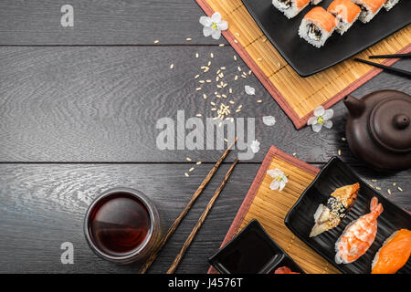 Sushi set Sushi, Sashimi, Nigiri und Maguro Rollen mit Teekanne und weiße Blumen auf dunklem Holz auf Teller serviert. Platz für Text. Stockfoto