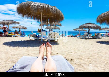 Beine und Füße ruhen auf einer der Sonnenliegen mit Sonnenschirmen im Hintergrund am Strand von Bahia Del Duque auf Teneriffa, Spanien Stockfoto