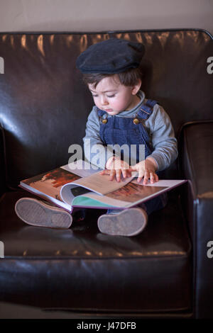 Süße kleine Junge mit braunen Haaren trägt ein blaues Denim Jeans Overalls und einem blauen Cord Cap hat ein Buch lesen in einem braunen Leder Stuhl Stockfoto