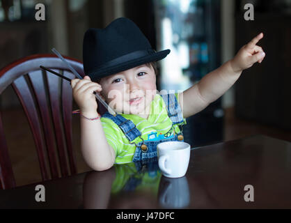 Süße kleine Junge mit braunen Haaren trägt einen schwarzen fedora Hut und Blue Denim jean Overalls ein gelbes Shirt mit weißem Porzellan Kaffee Tasse Tee Stockfoto