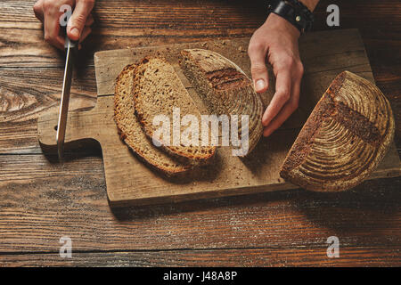 Geschnittenes Brot auf einem braunen Holzbrett mit Mannes Hand hält ein Messer flach zu legen Stockfoto