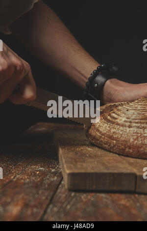Der Mann schneidet frisches Bauernbrot auf einem hölzernen Hintergrund Stockfoto