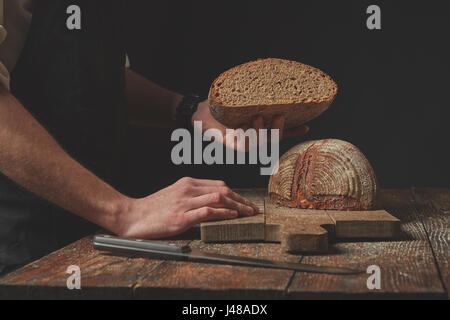 Baker hält die Hälfte frisch gebackenes Bio-Brot auf einem dunklen Hintergrund Stockfoto