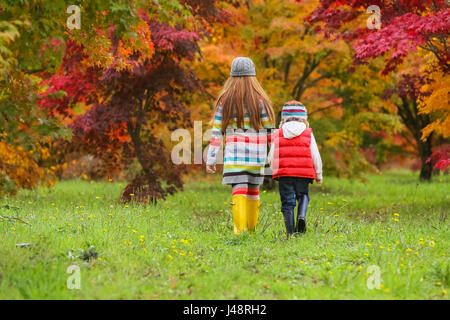 Ein kleiner Junge und ein Mädchen Gummistiefel und bunte Kleidung tragen Fuß über ein Feld in Richtung Bäume in leuchtenden Herbstfarben Stockfoto